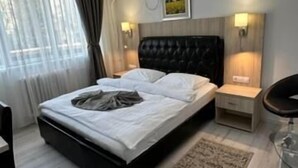 A modern bedroom with a black upholstered headboard, a large bed, two bedside tables with lamps, a small bedside table, a chair, and a view of the outdoors through sheer curtains. - Bulevard Prestige Hotel (Slatina)