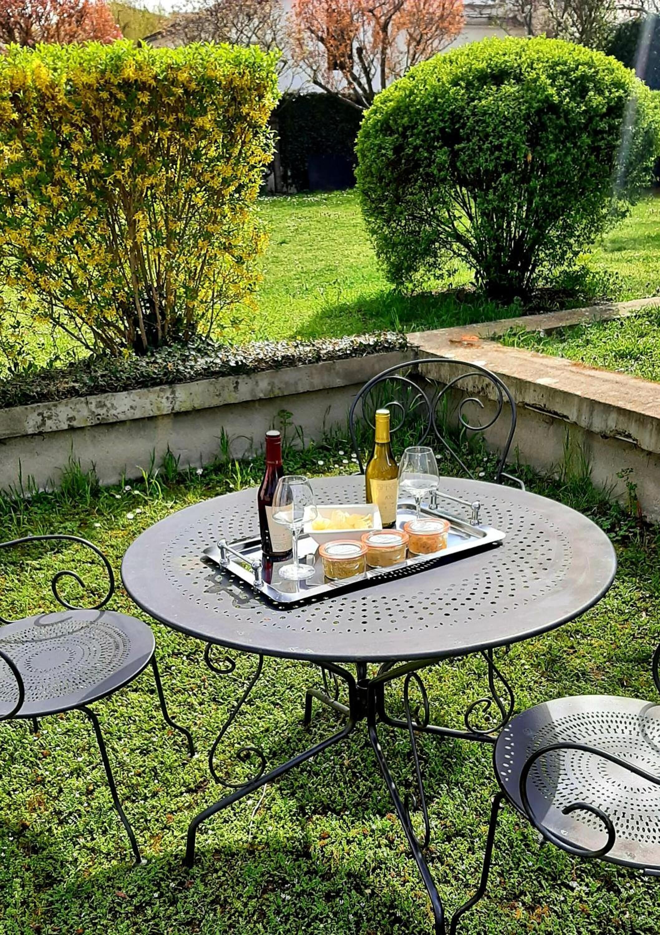 A round metal table with a tray of food and drinks, surrounded by metal chairs, set on a grassy lawn with bushes in the background.