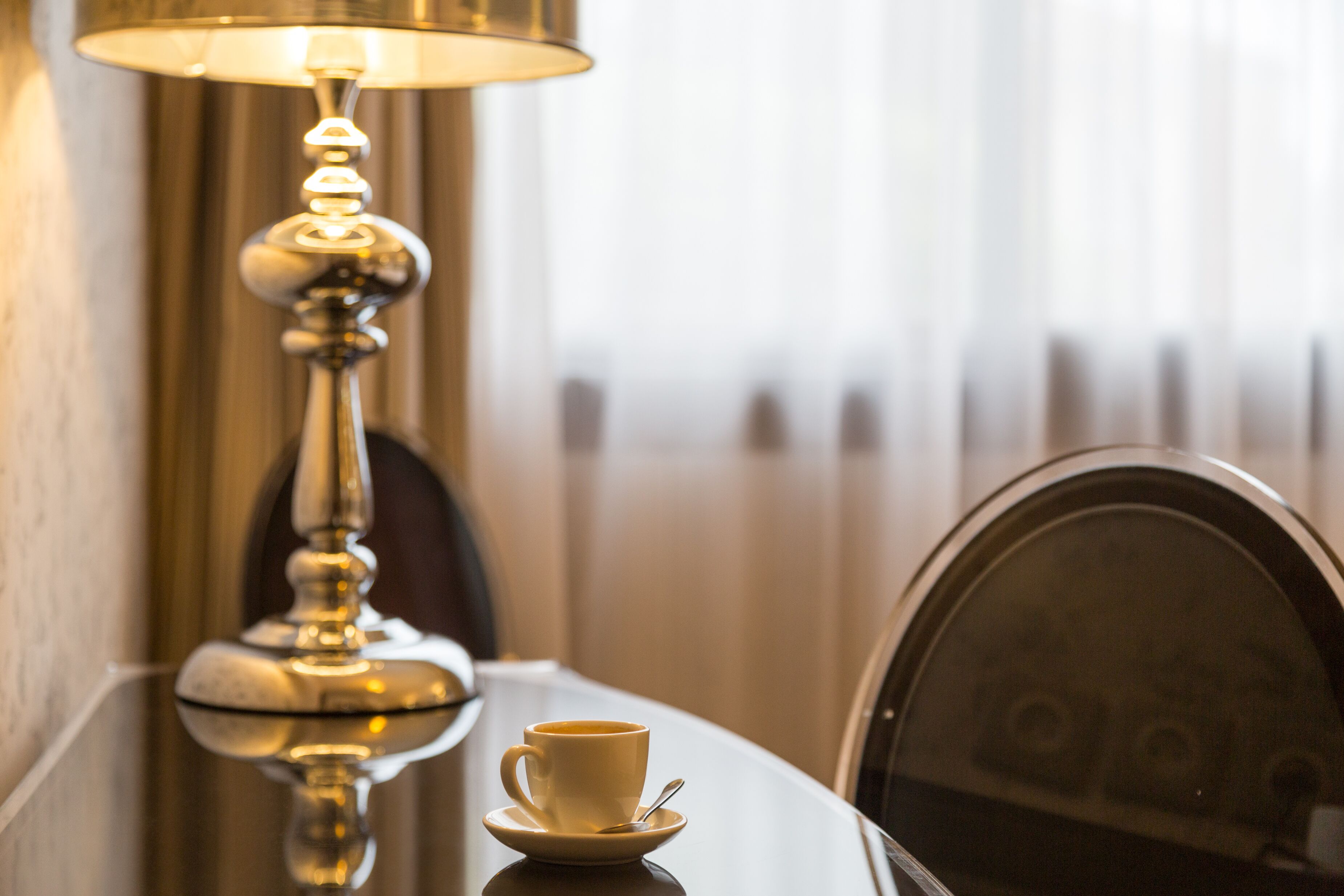 A table with a cup of coffee and a saucer, a lamp, and a chair with a reflective surface.