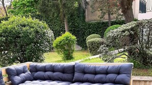 Un canapé modulable posé sur des palettes en bois, un foyer circulaire entouré de bûches et une pelouse verdoyante agrémentée d’arbres et d’arbustes.