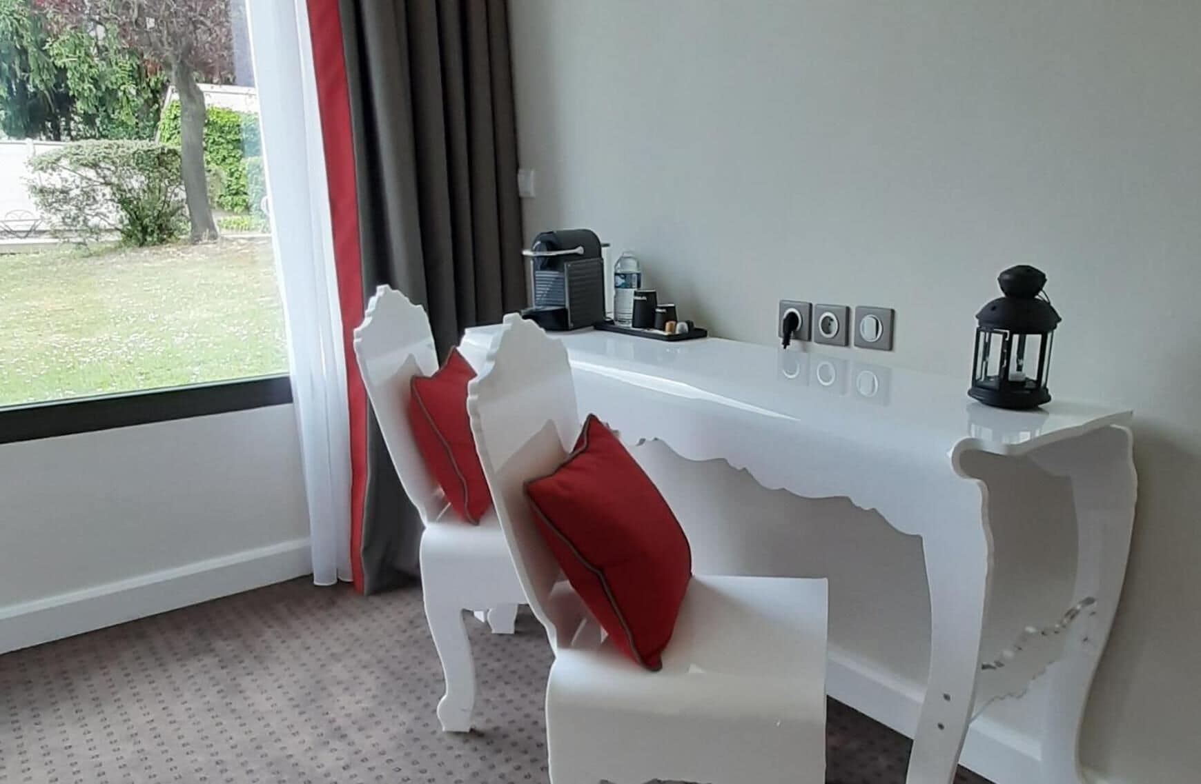 A modern room with a white, curved desk, red cushions, a coffee machine, and a view of a garden.