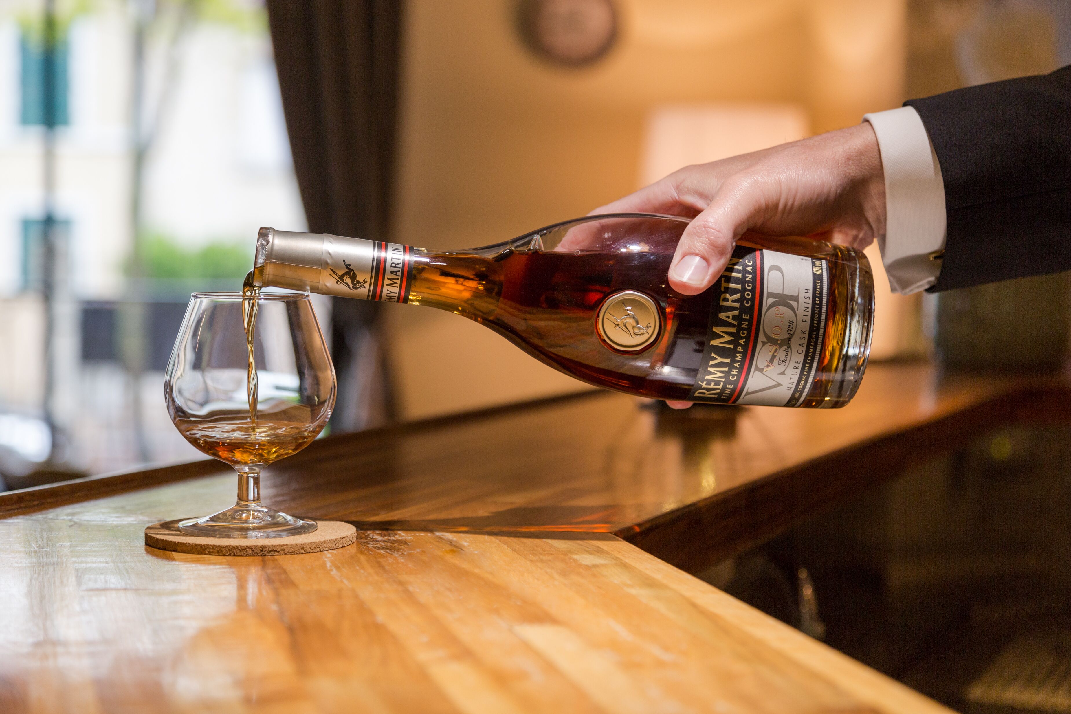 Une main verse le contenu d’une bouteille de whisky dans un verre, sur un comptoir en bois.