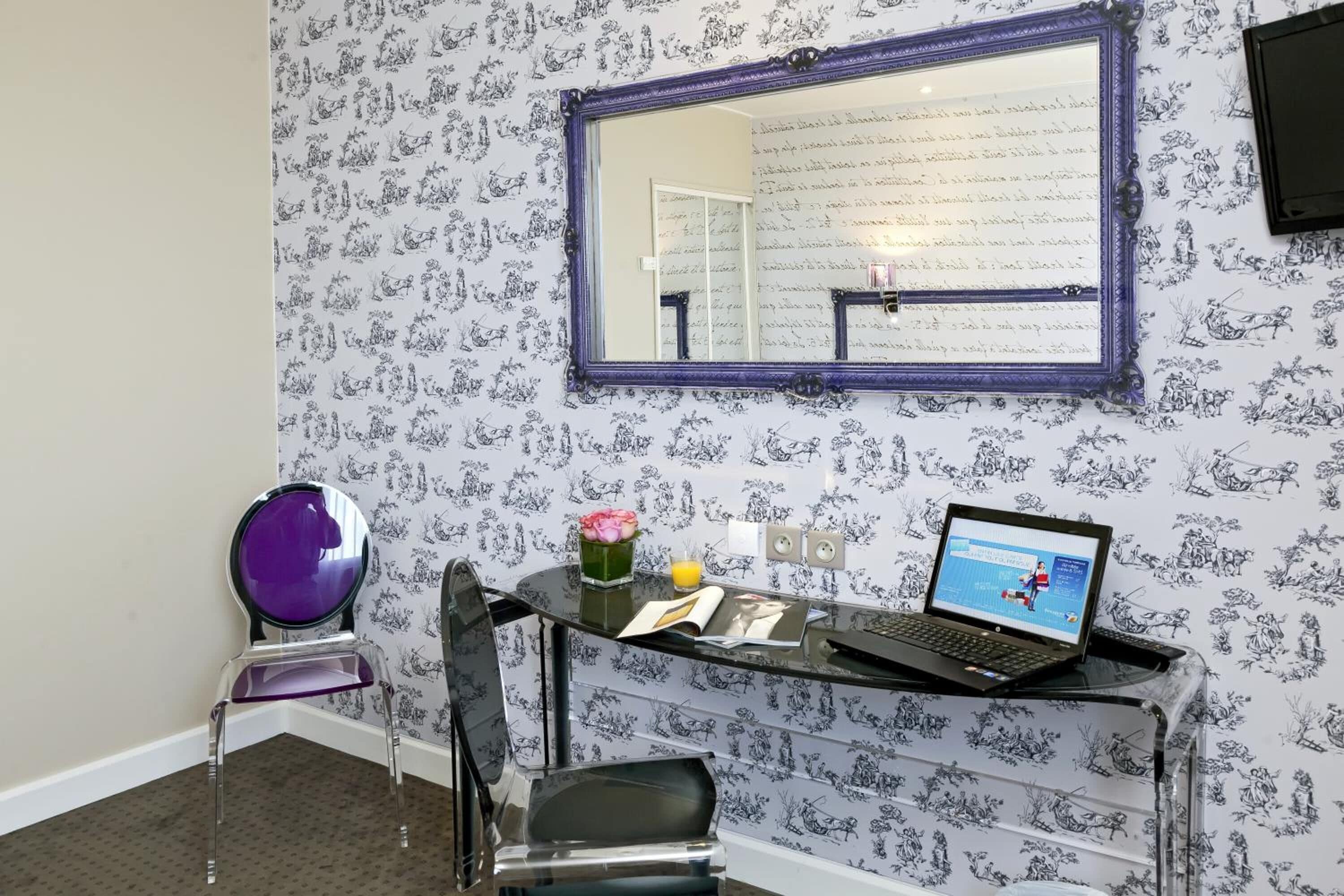 A room with a glass desk, a laptop, a vase with flowers, and a mirror on the wall.