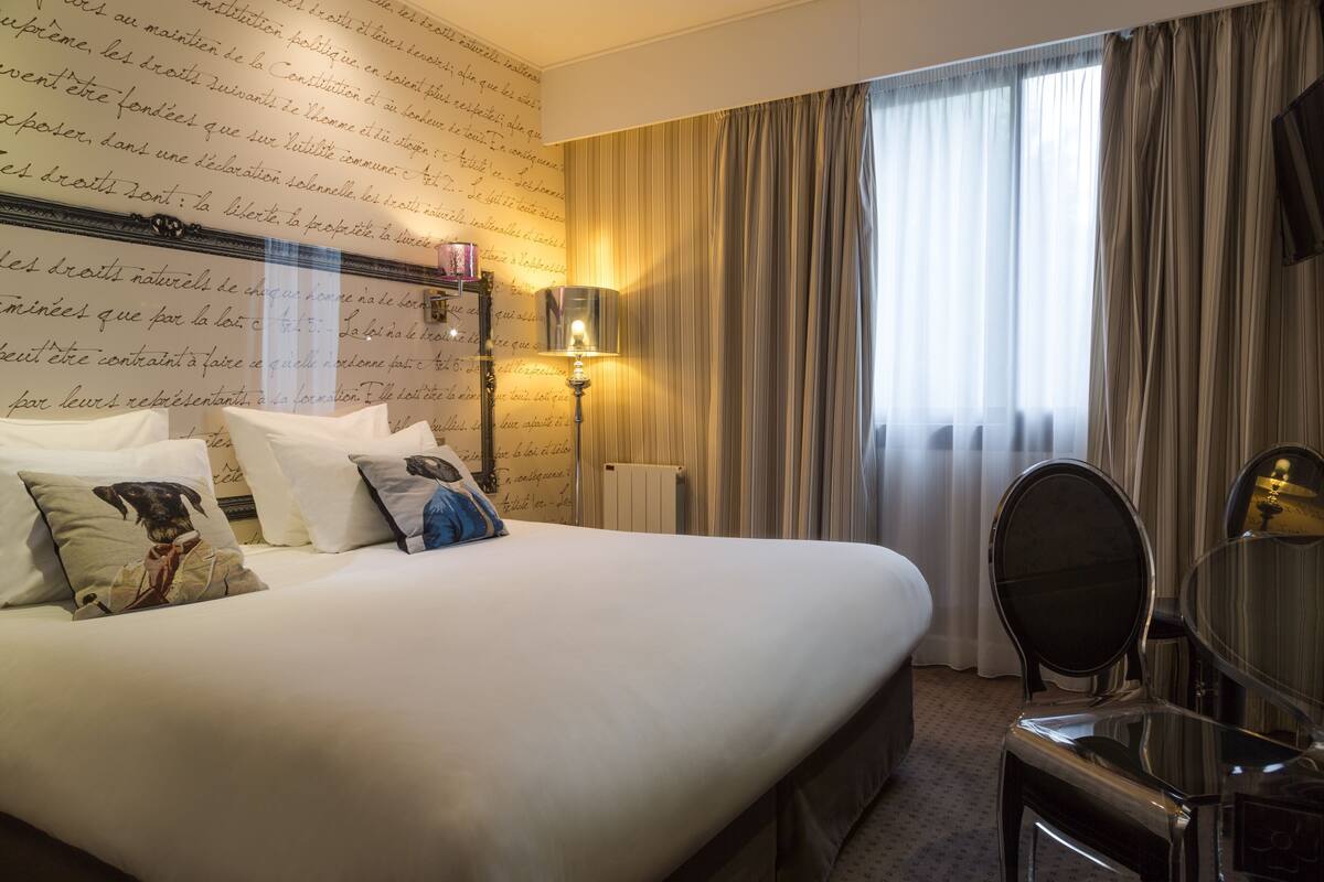 Een hotelkamer met een groot bed, een nachtkastje met een lamp, een stoel en een raam met gordijnen.