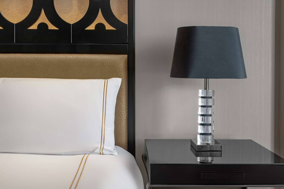 Ein ordentlich bezogenes Bett mit weißer Bettwäsche und goldenen Akzenten, eine moderne Lampe auf einem Nachttisch und ein gemustertes Kopfteil.