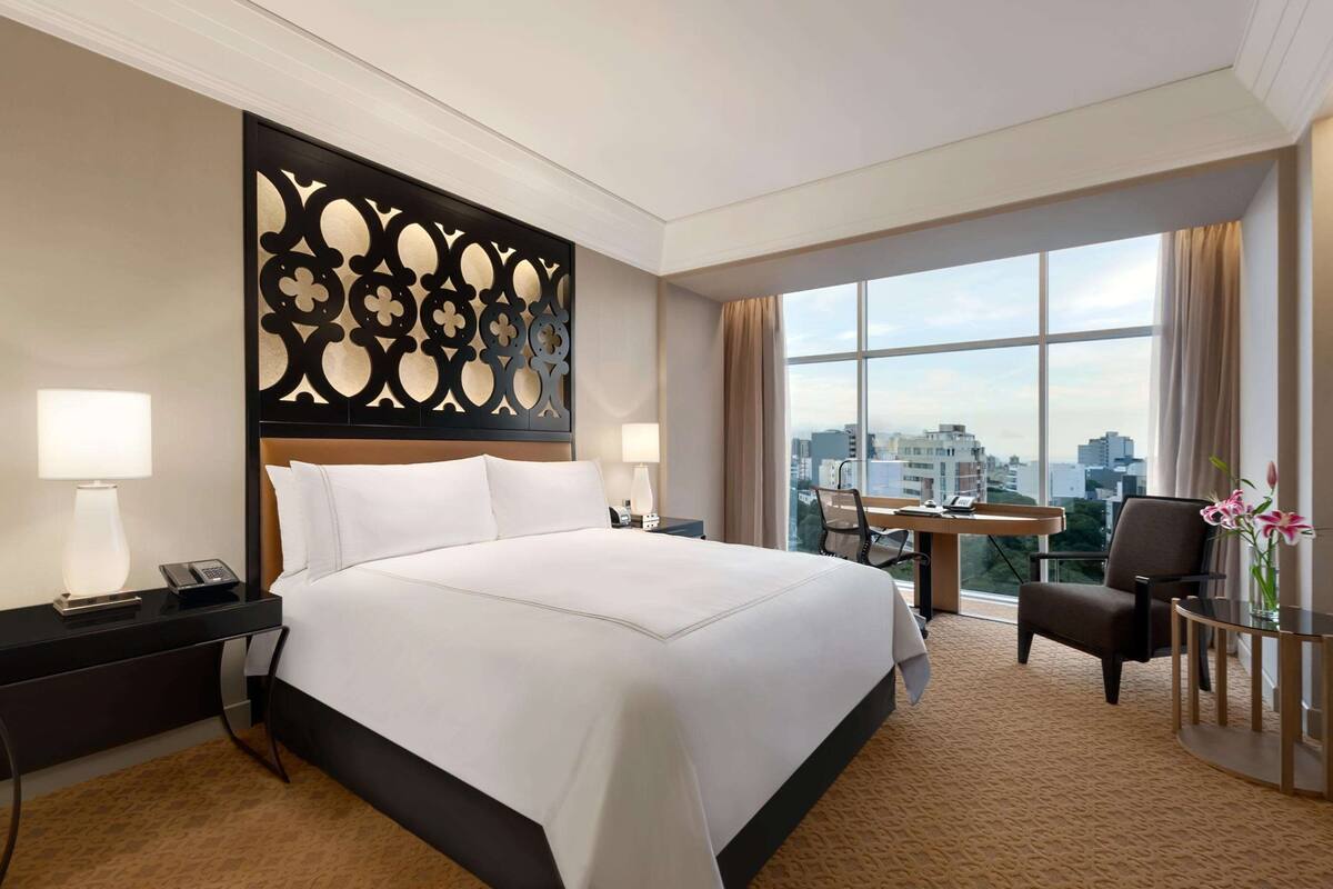 Ein Hotelzimmer mit einem großen Bett, einem Schreibtisch, einem Sessel und Blick auf die Stadt durch große Fenster.