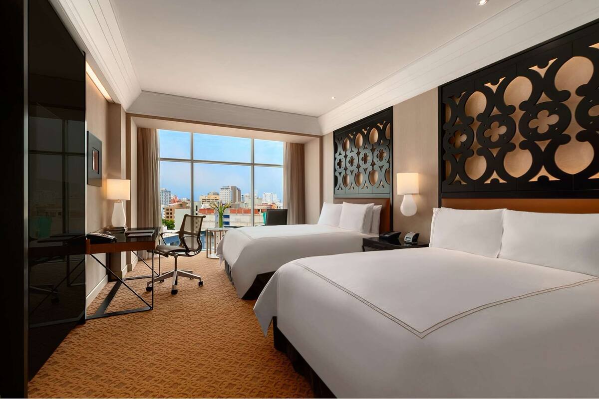 Ein Hotelzimmer mit zwei Betten, einem Schreibtisch mit Stuhl, einem großen Fenster und einer Zierleiste an der Wand.