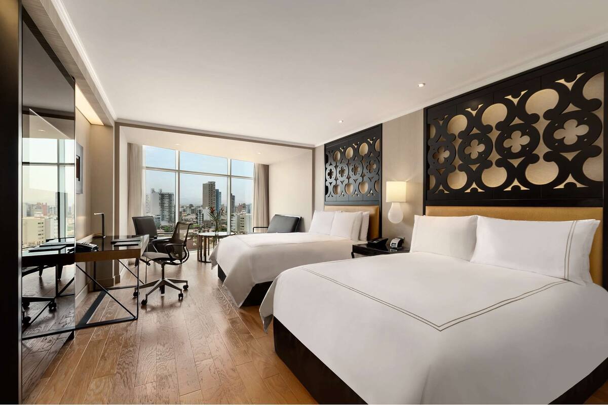 Ein Hotelzimmer mit zwei Betten, einem Schreibtisch und einem großen Fenster mit Blick auf die Stadt.