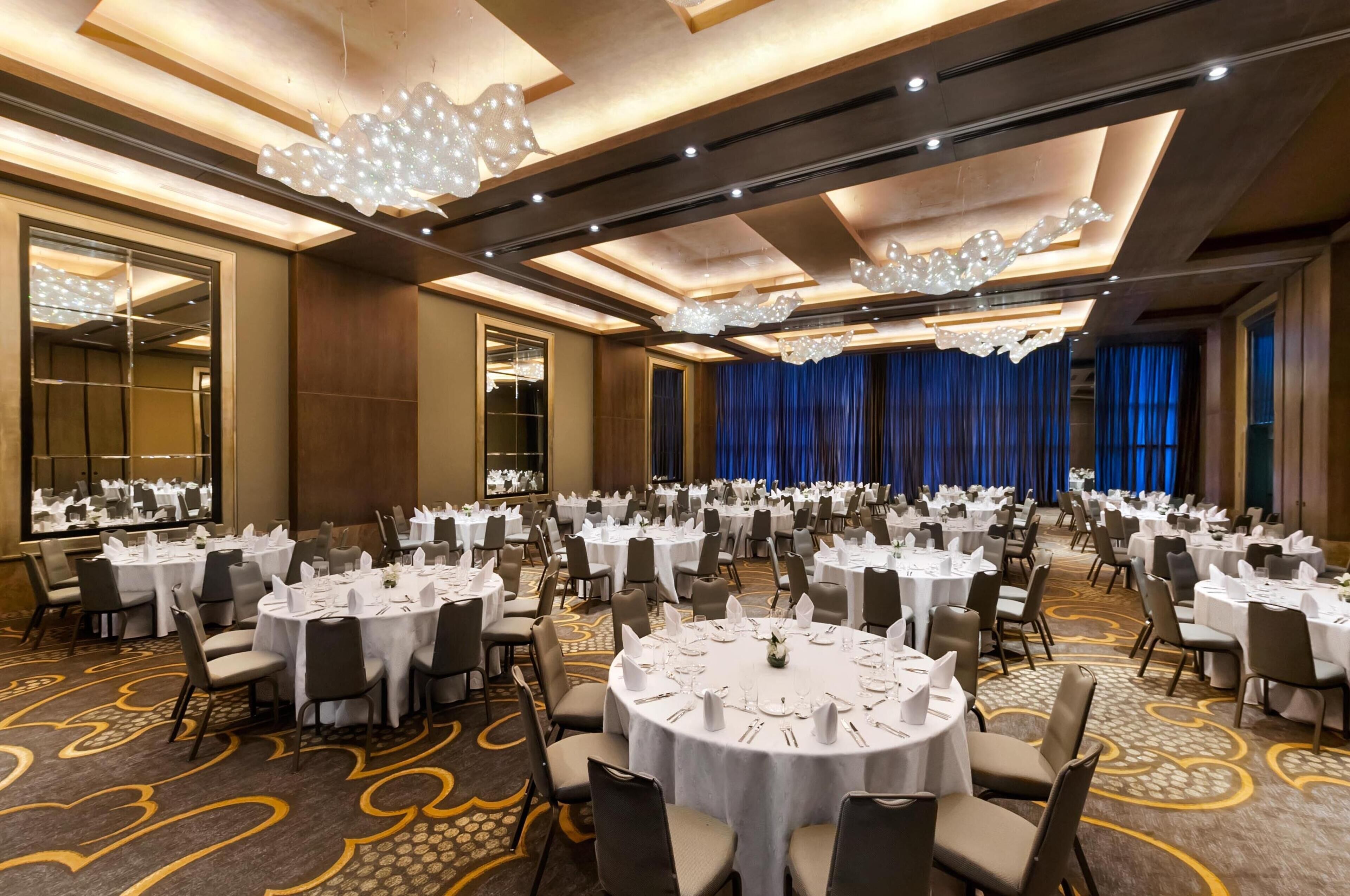 Un salón de banquetes con mesas redondas dispuestas para comer, un escenario con cortinas y una pared reflectante.