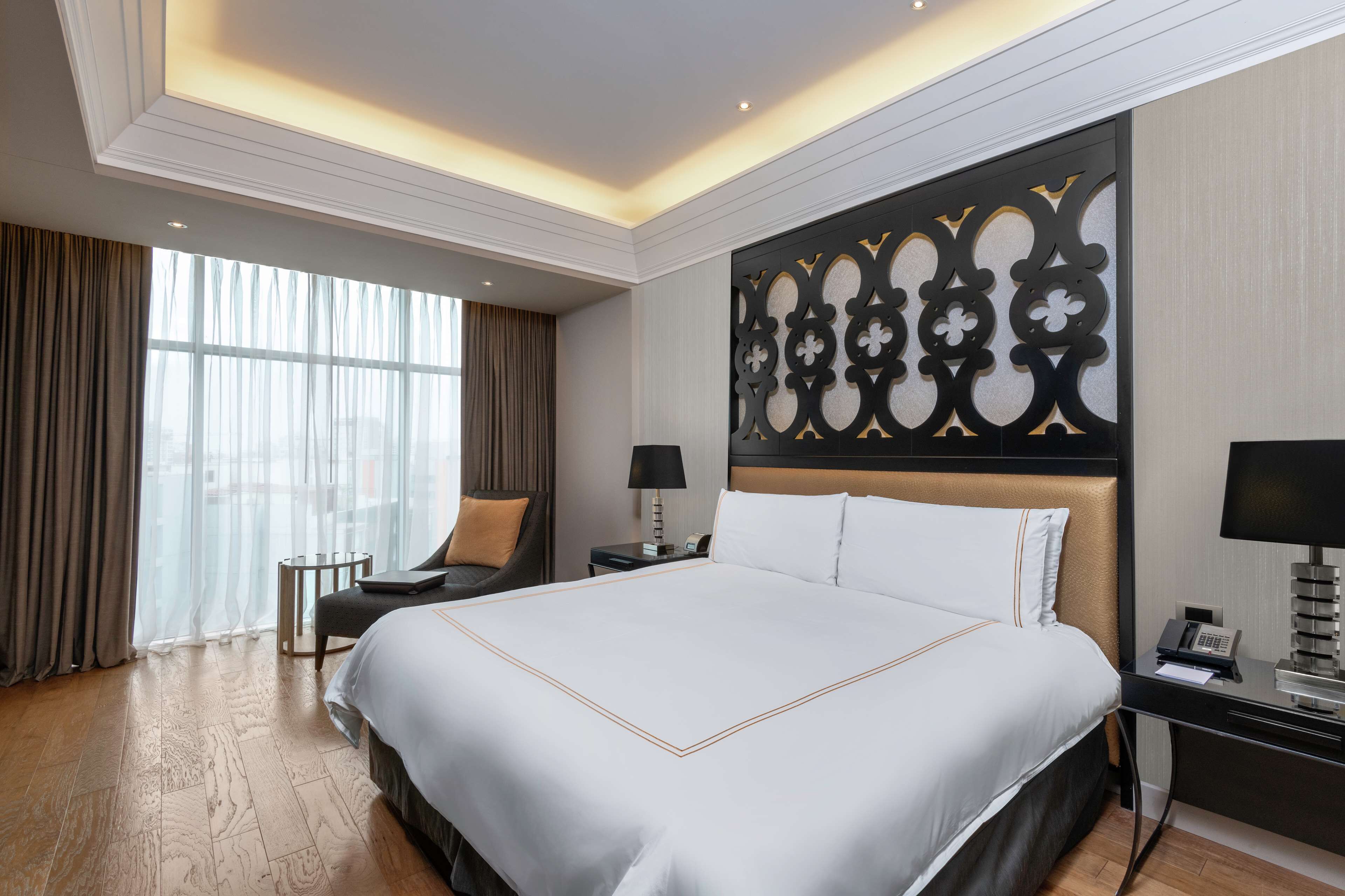 A hotel room with a large bed, bedside tables, a desk, a chair, and a decorative wall panel.