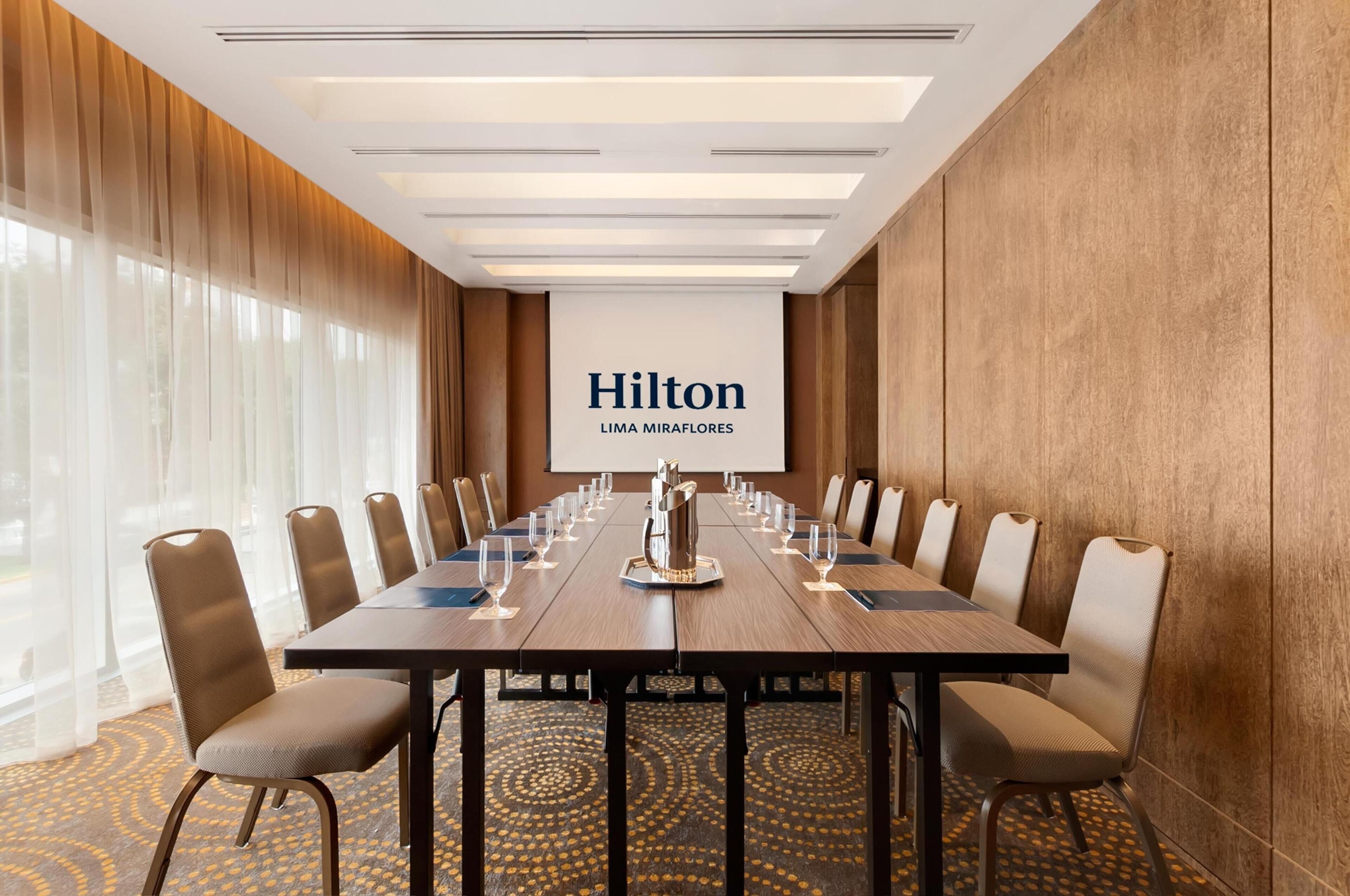 Un salón de conferencias con una larga mesa, sillas y una pantalla que muestra 'Hilton LIMA MIRAFLORES'.