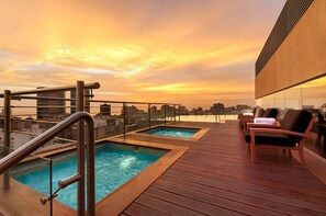 Zona de piscina en la azotea con cubierta de madera y dos piscinas, rodeada de sillones de descanso y vistas a la ciudad.