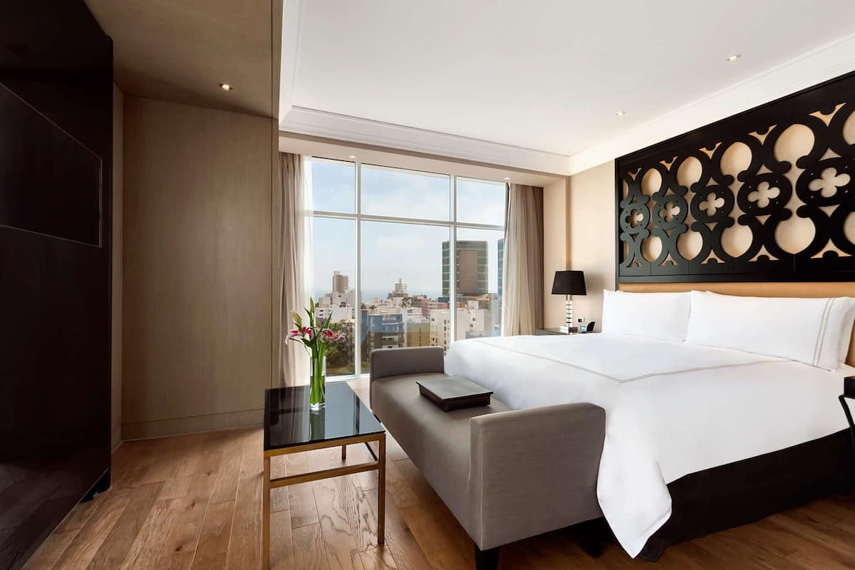 Ein Hotelzimmer mit einem großen Bett, einem Schreibtisch, einem Sessel und Blick auf die Stadt, sowie einer dekorativen Wandverkleidung.