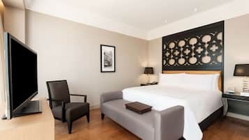 A hotel room with a large bed, a flat-screen TV, a desk with a lamp, a chair, and a small table.