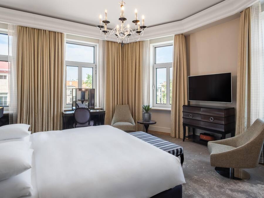 A hotel room with a large bed, a TV, a desk with a chair, and a chair with a plant.