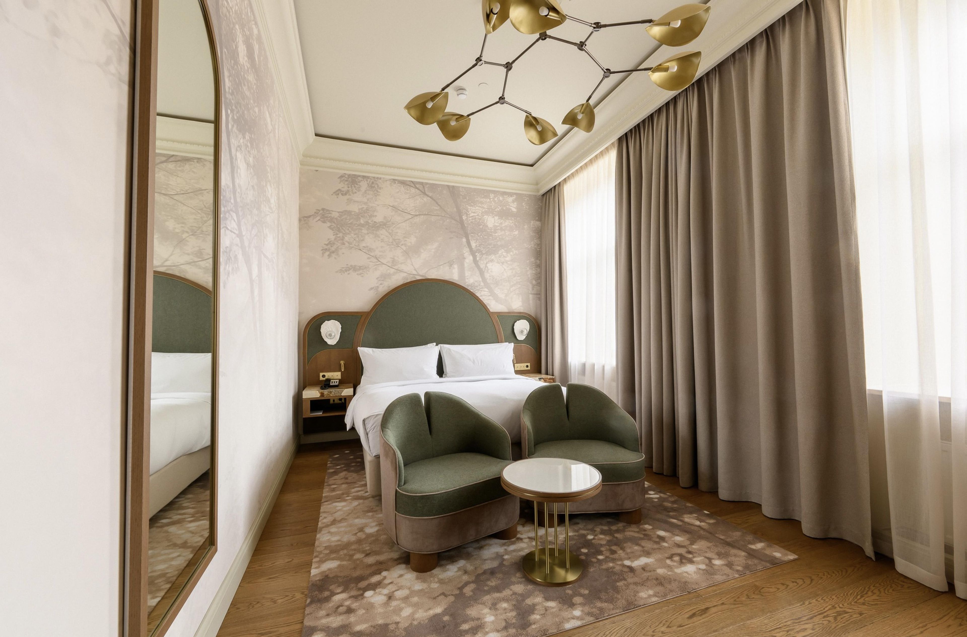 Une chambre d’hôtel avec un lit, deux fauteuils, une petite table ronde, un miroir et un lustre.