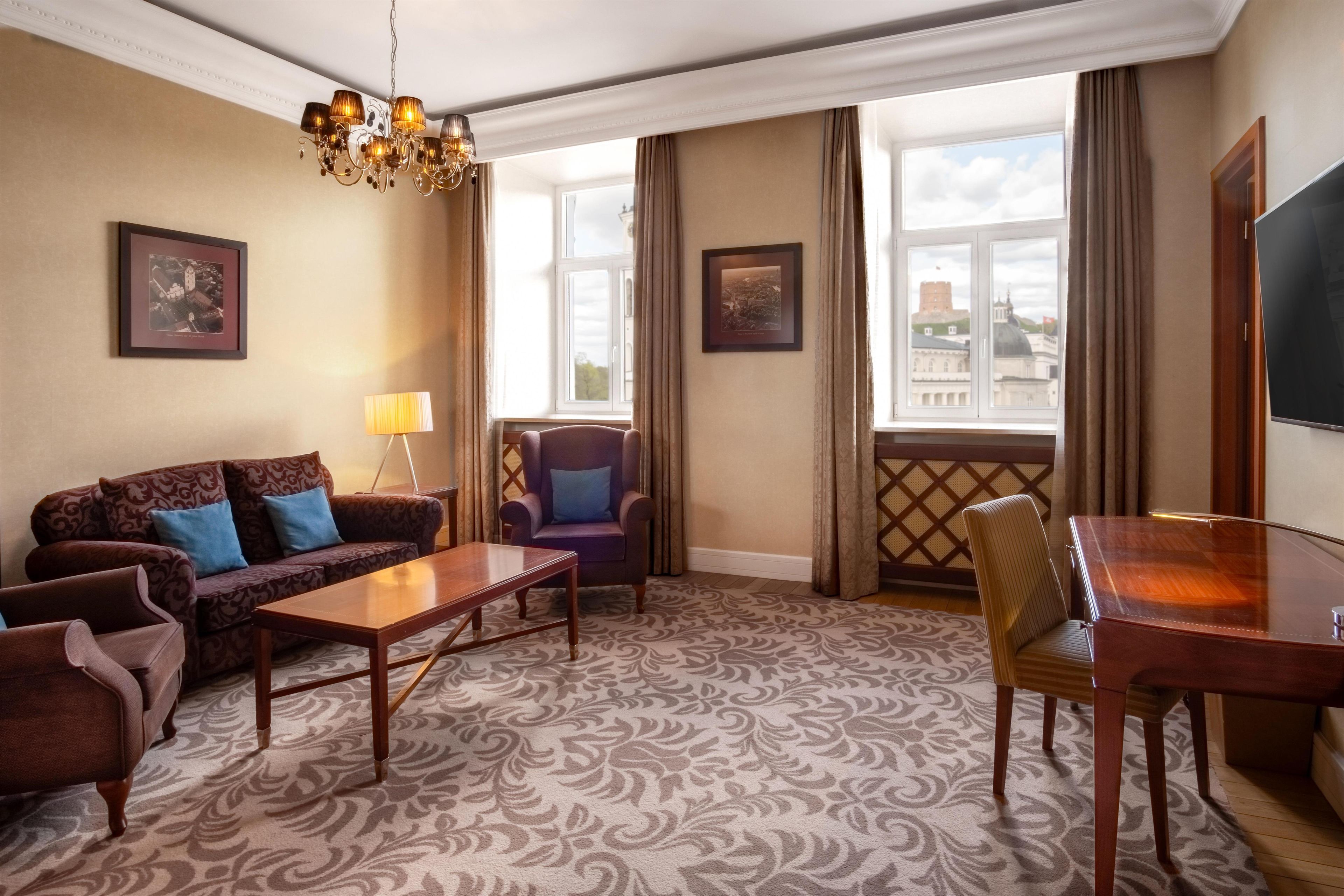 A hotel room with a patterned carpet, a sofa set, a dining table, a chair, a lamp, a chandelier, and two framed pictures on the walls.