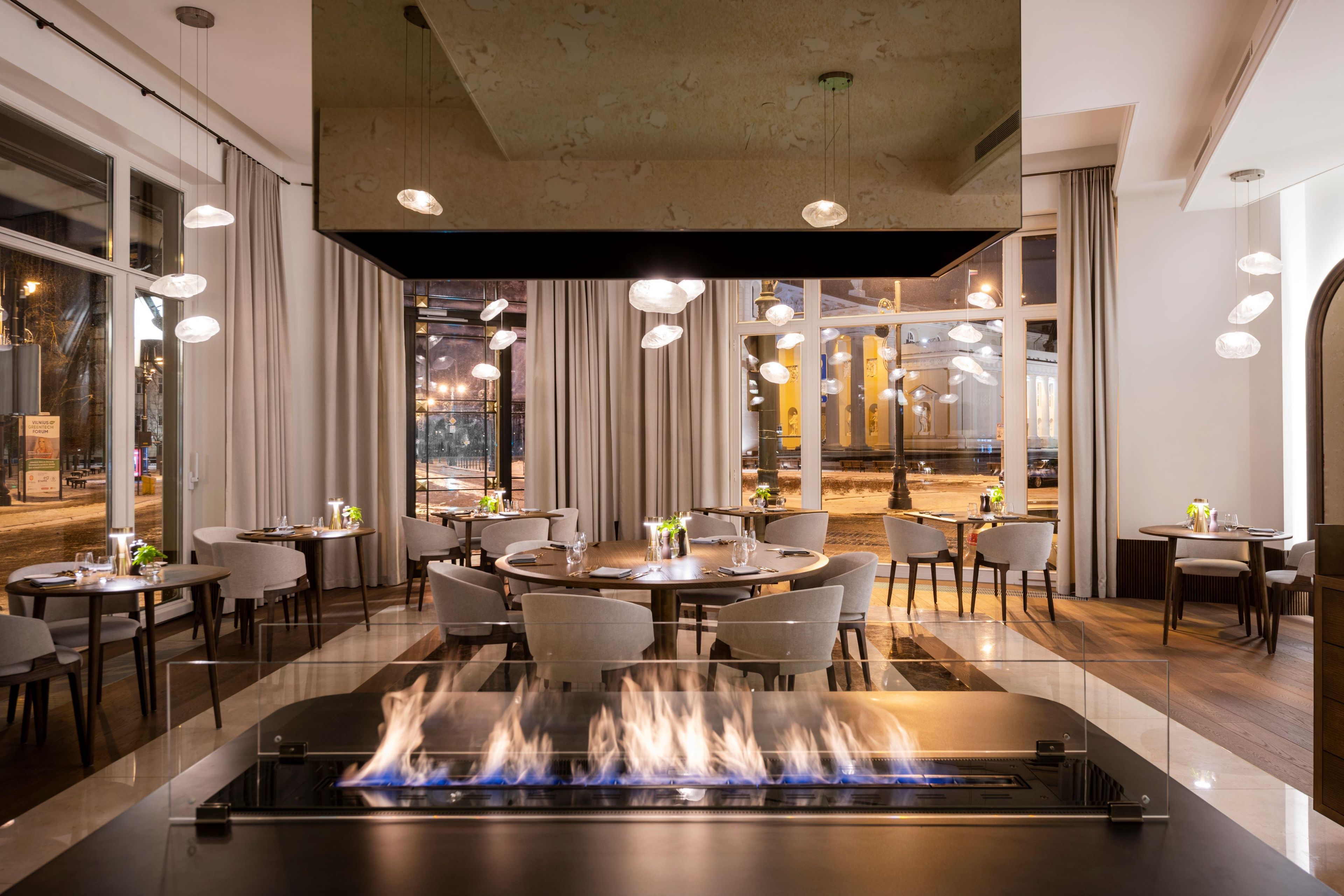 A restaurant with round tables, chairs, and a central fire feature.