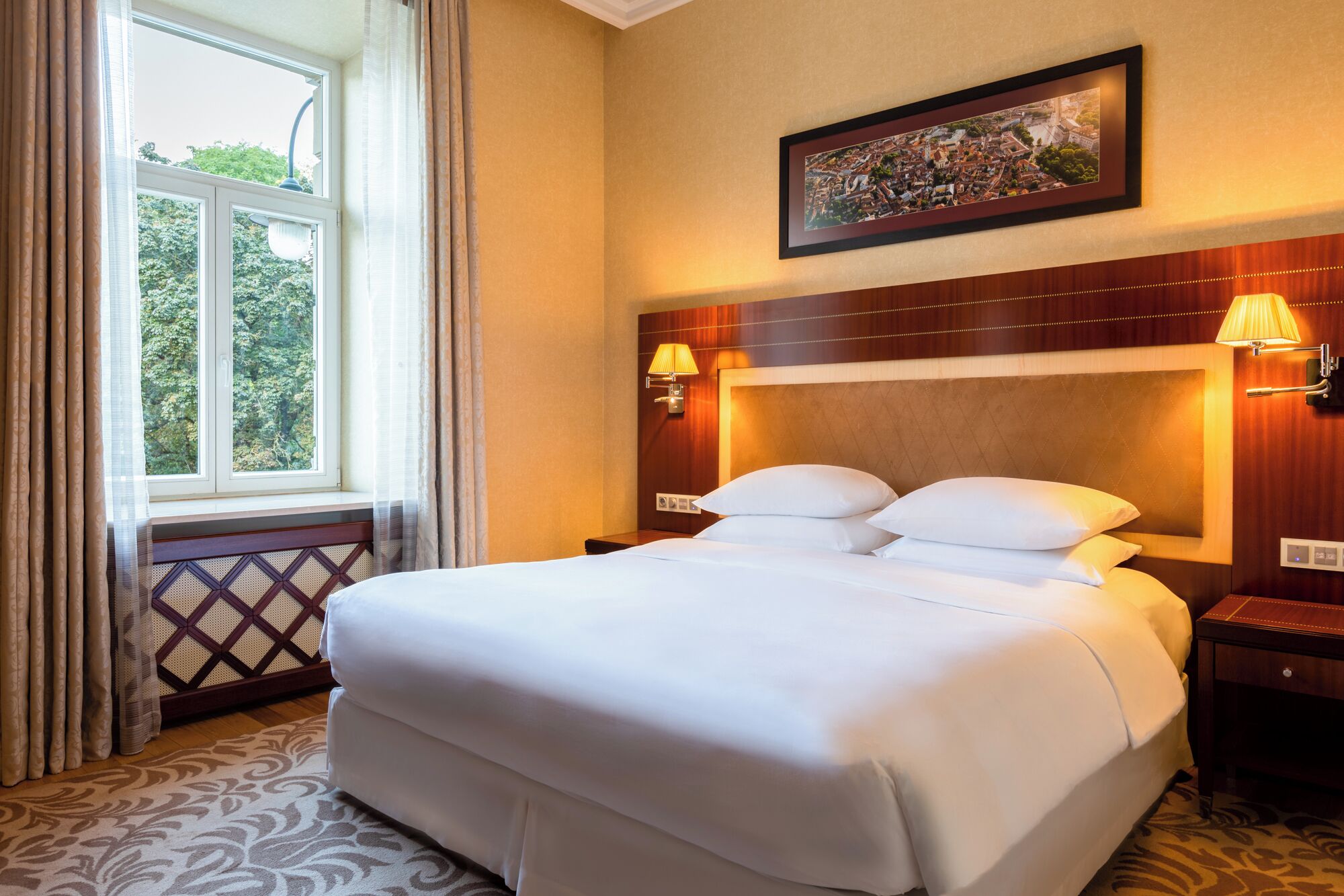 A hotel room with a large bed, two bedside lamps, a framed picture on the wall, and a window with curtains.