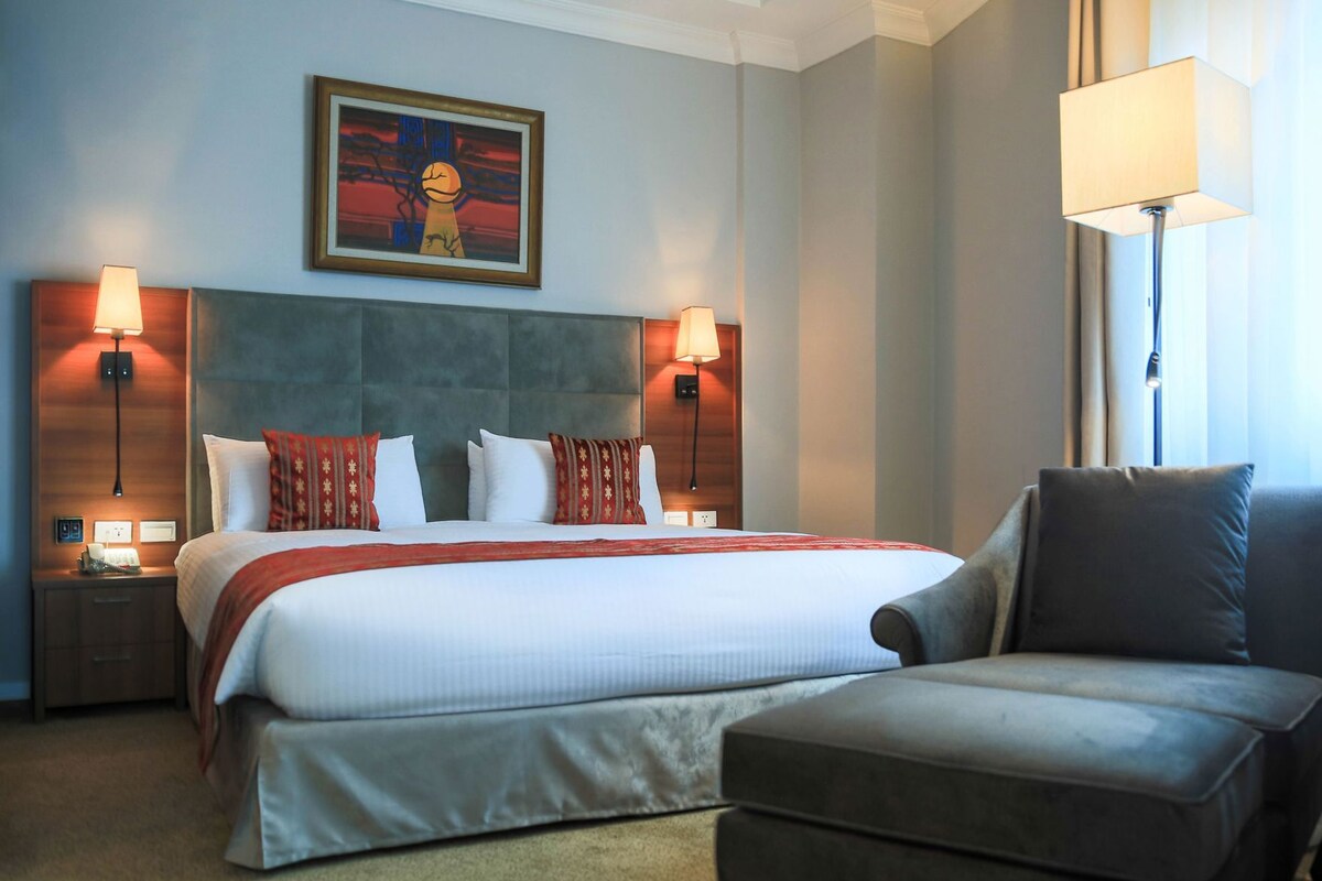 Habitación de hotel con una cama grande, dos lámparas de noche, una obra enmarcada sobre la cama y un sofá.
