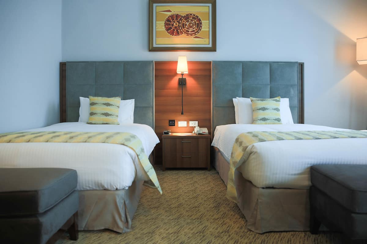A hotel room with two beds, a nightstand with a lamp, and a wall-mounted artwork.