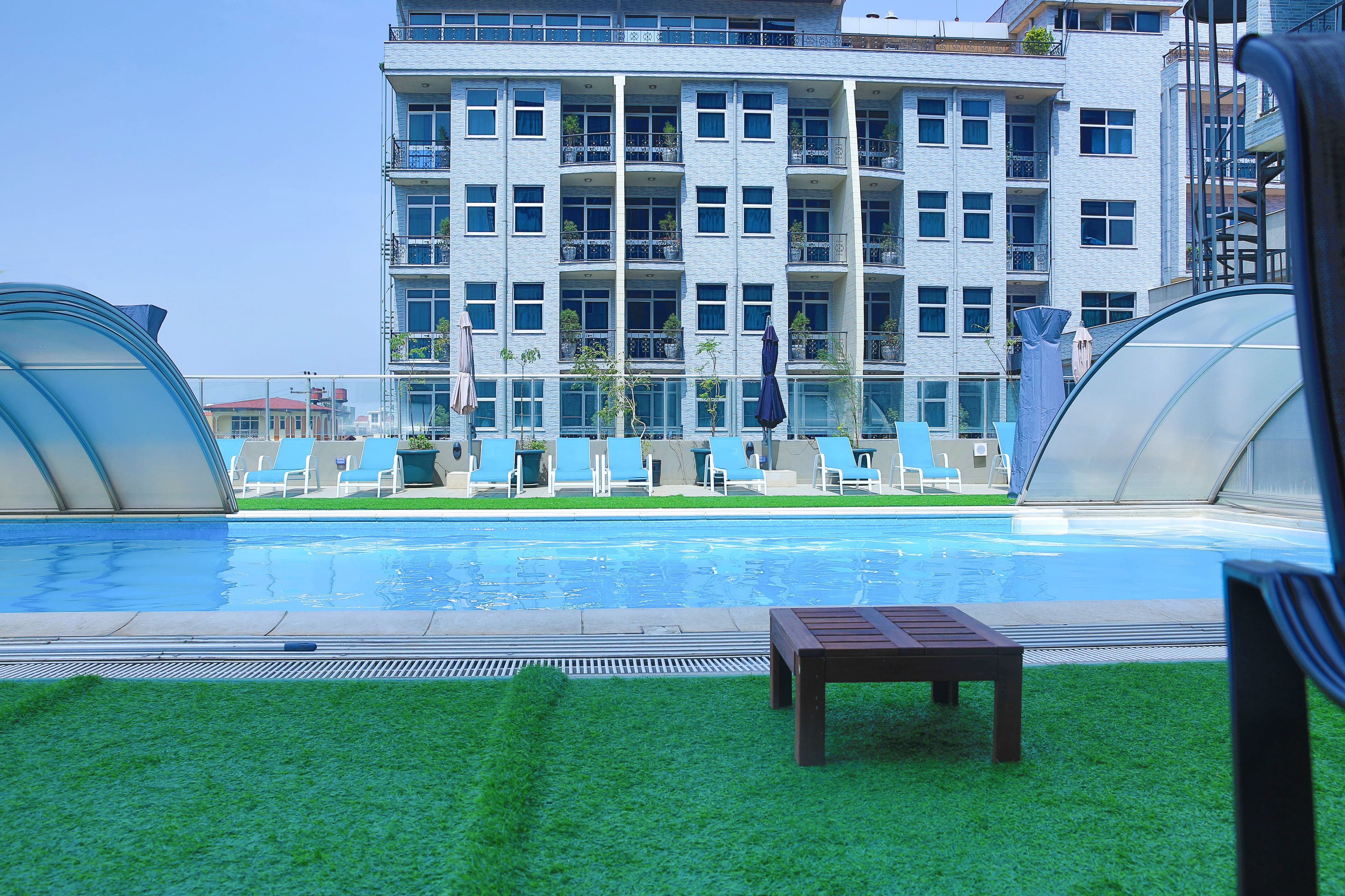 A swimming pool with lounge chairs and a wooden bench, surrounded by a building with multiple balconies and windows.