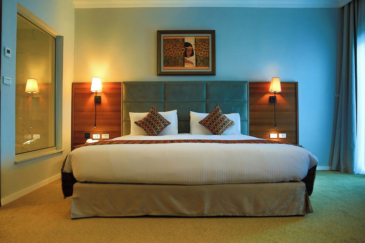 Una habitación de hotel con una cama grande, dos lámparas de noche, un cabecero y una obra de arte enmarcada sobre la cama.