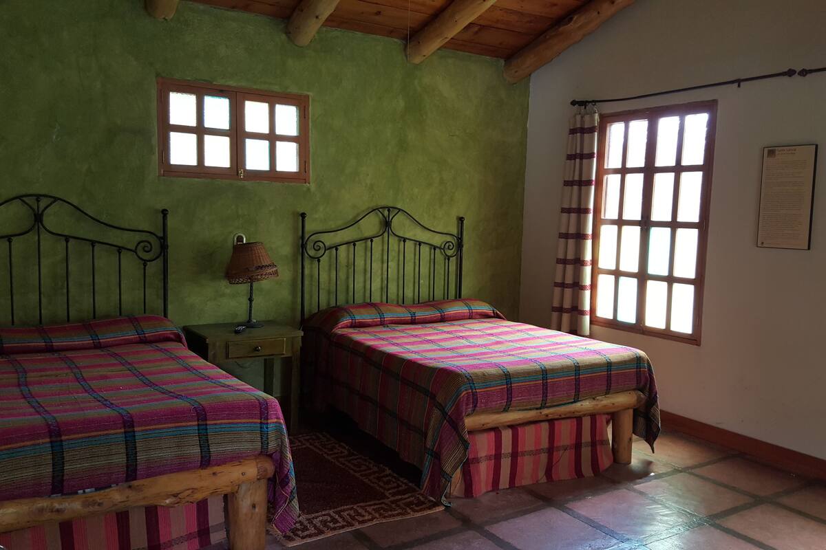 A room with two beds, a small table with a lamp, and a window with curtains.