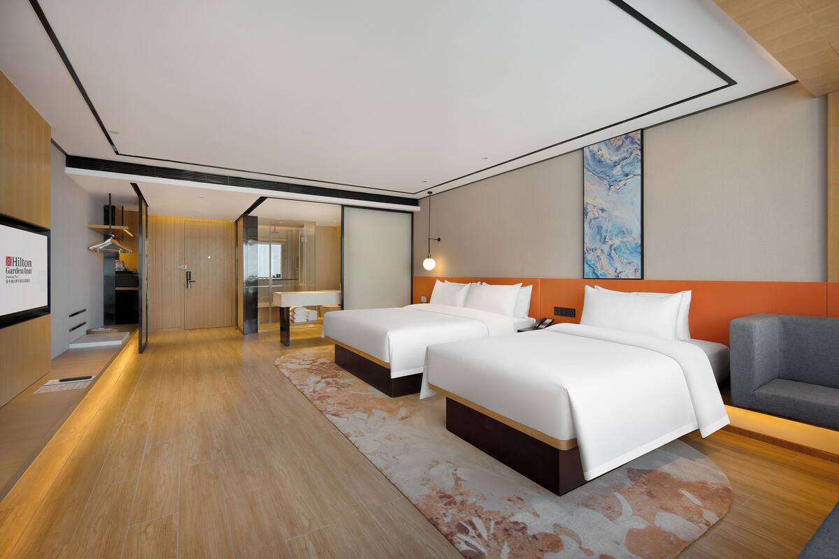 A modern hotel room with two beds, a sofa, a desk, and a large window.