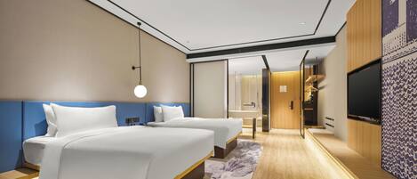 A modern hotel room with two beds, a large mirror, a TV, and a bathroom visible through a glass door.