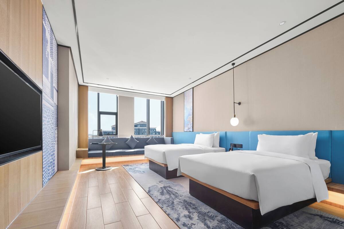 A modern hotel room with two beds, a large window, a flat-screen TV, and a city view.