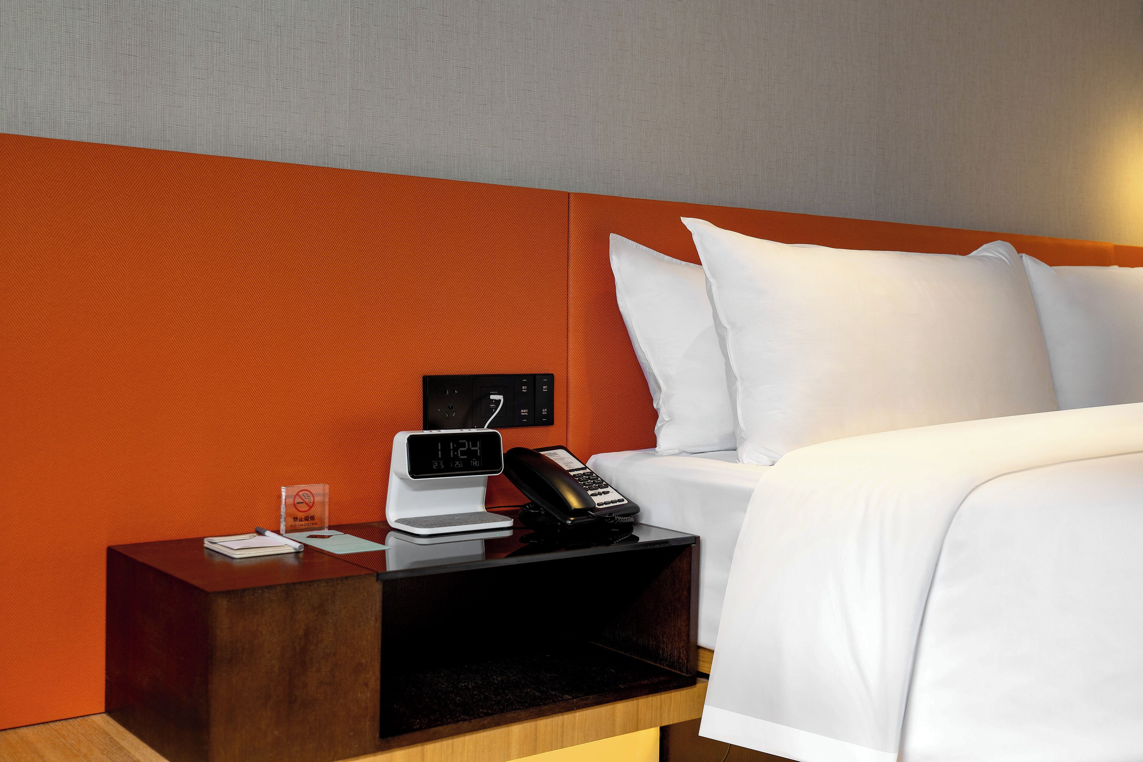 A hotel room with a bed, white bedding, a wooden nightstand with a clock, a phone, and a small table with a notepad and pen.