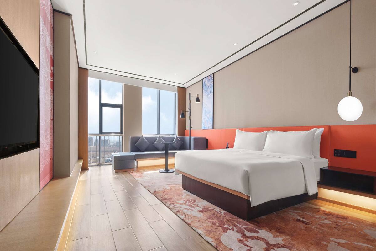 A modern hotel room with a large bed, a sofa, a small table, and a view of the city through large windows.