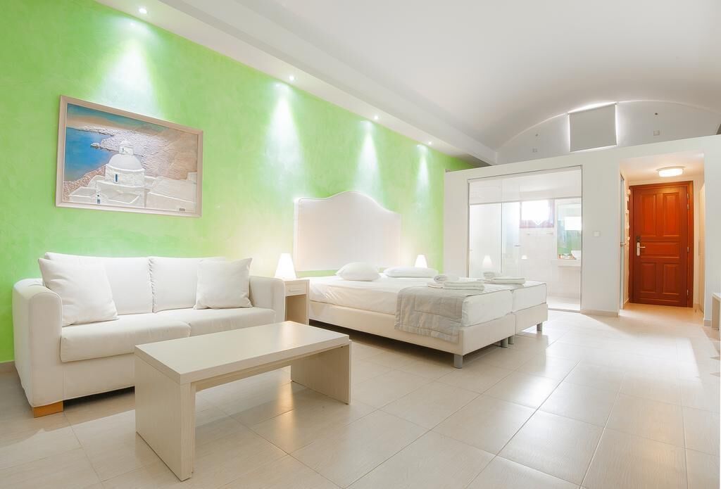A modern hotel room with a green accent wall, a white sofa, a bed with white linens, and a painting of a white building.