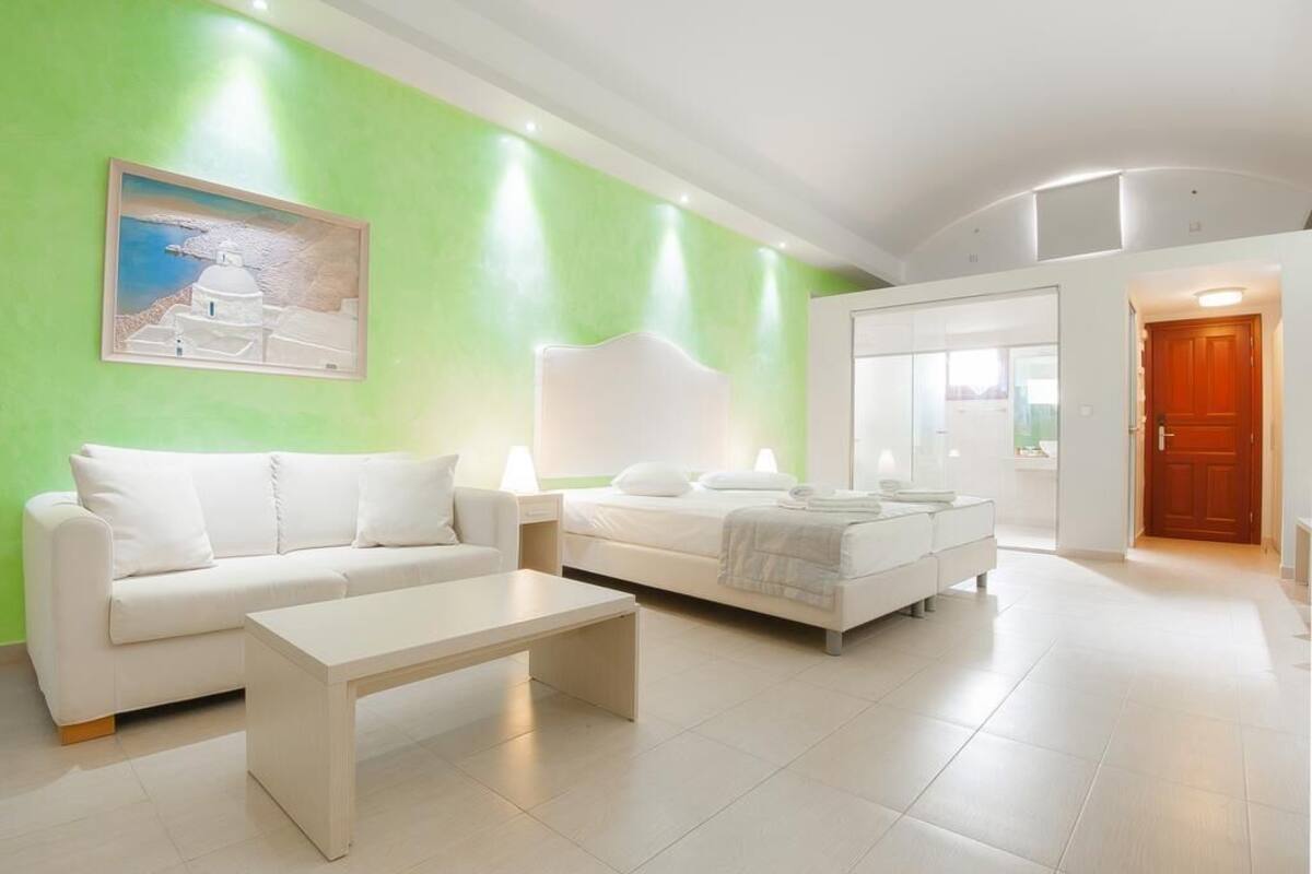 A modern hotel room with a green accent wall, a white sofa, a bed with white linens, and a painting of a white building.
