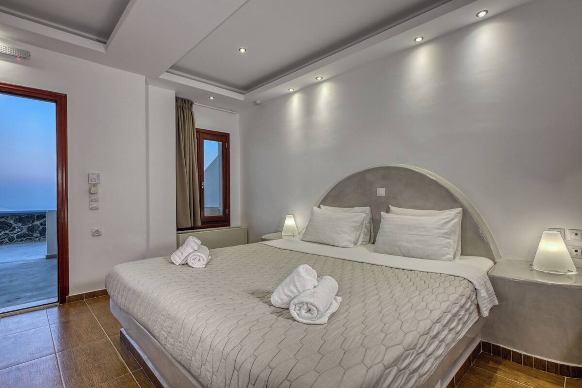 Une chambre d’hôtel moderne avec un grand lit, une table de chevet avec une lampe et une vue sur la mer, visible à travers une porte ouverte.