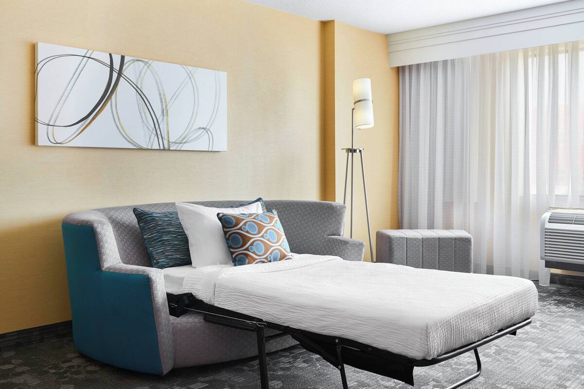 A modern hotel room with a sofa bed, a standing lamp, and a large abstract painting on the wall.