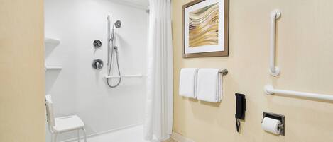 A bathroom with a shower, a toilet, a chair, a towel rack, a handrail, and a phone.