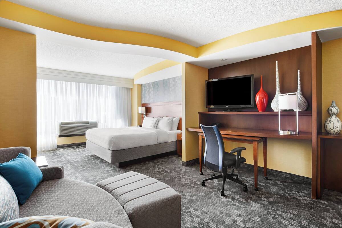 A hotel room with a bed, a desk with a chair, a television, and a large window with curtains.