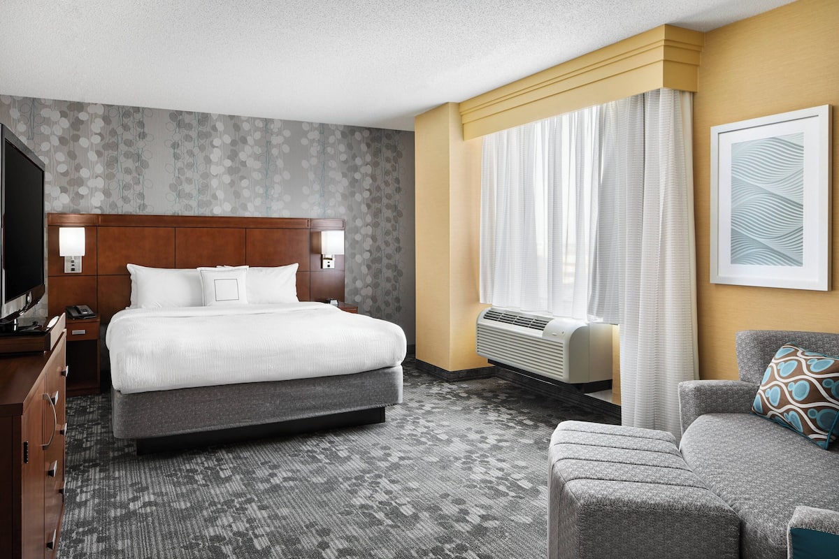A hotel room with a large bed, a desk with a lamp, a chair, a TV, and a window with curtains.