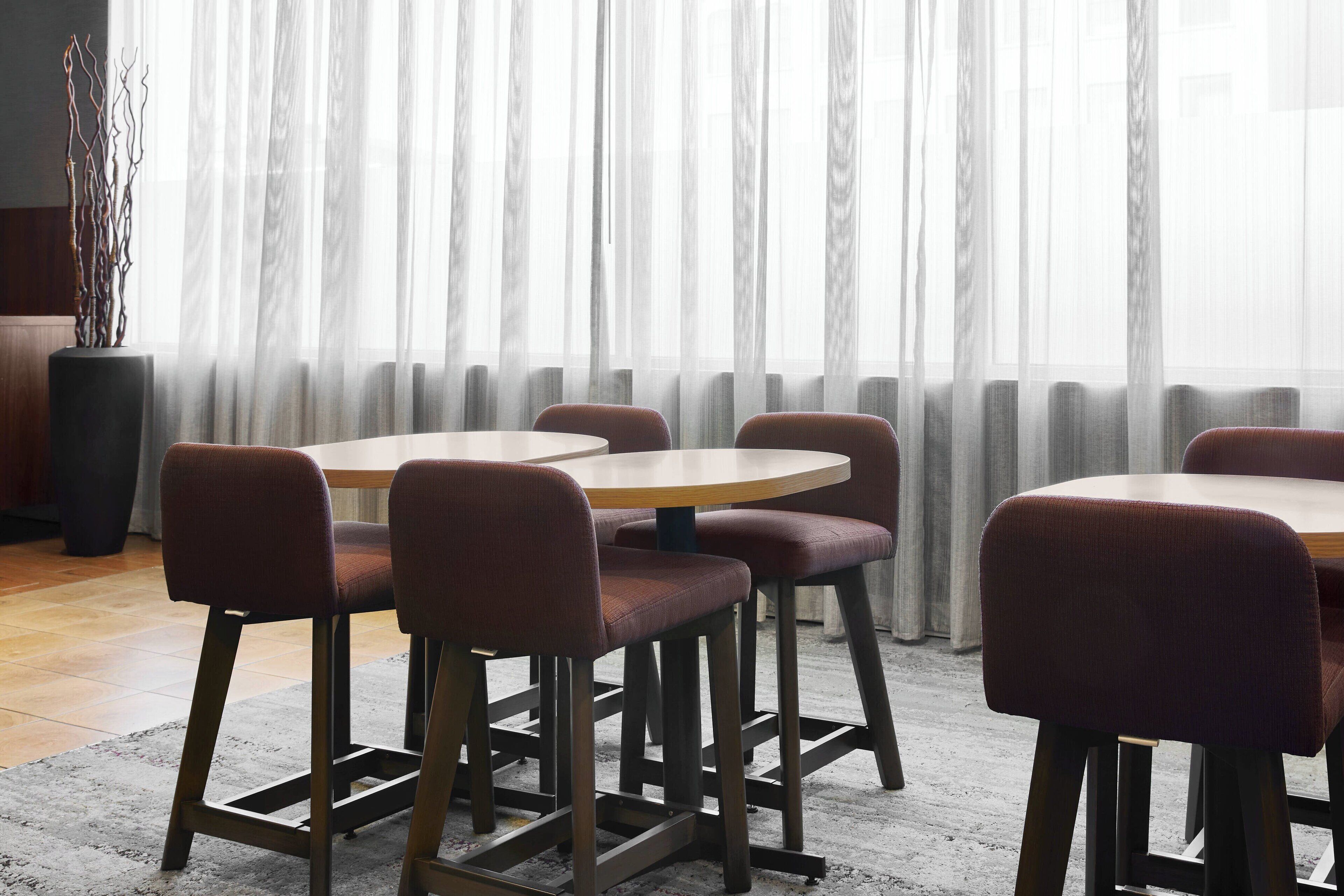 Un espace repas avec des tables rondes et des chaises rembourrées, une grande plante en pot et des rideaux légers.