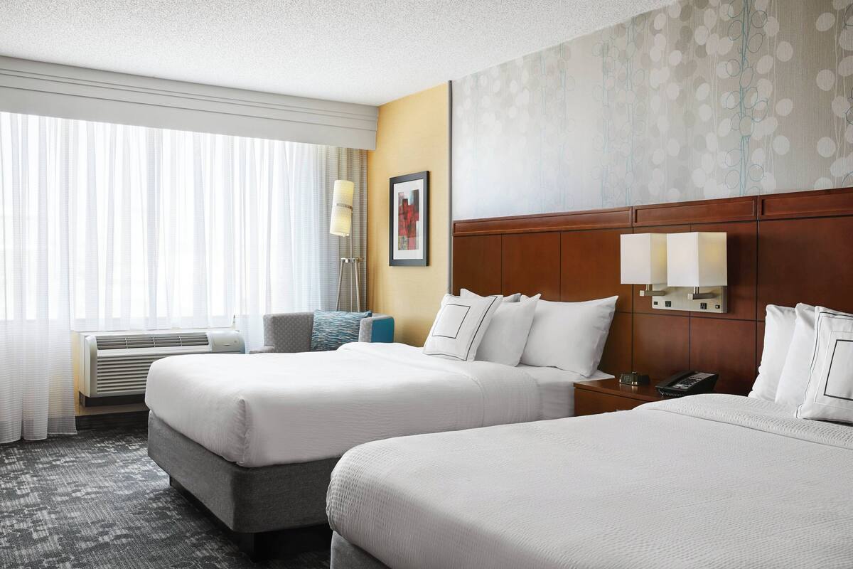 A hotel room with two beds, a large window with curtains, a wall-mounted lamp, and a framed picture.