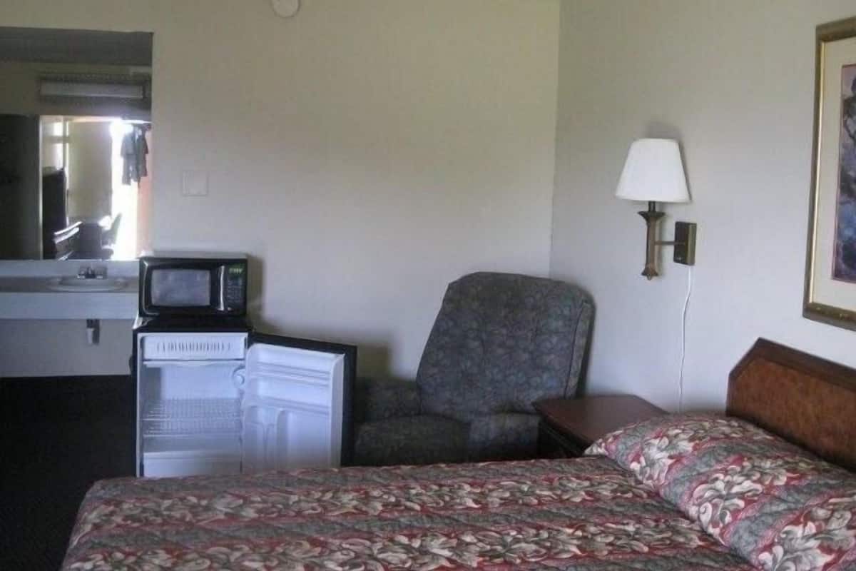A hotel room with a bed, a chair, a desk with a computer, a lamp, and a painting on the wall.
