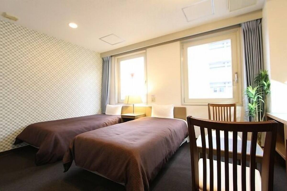 A hotel room with two beds, a desk with a chair, a window with curtains, and a plant.