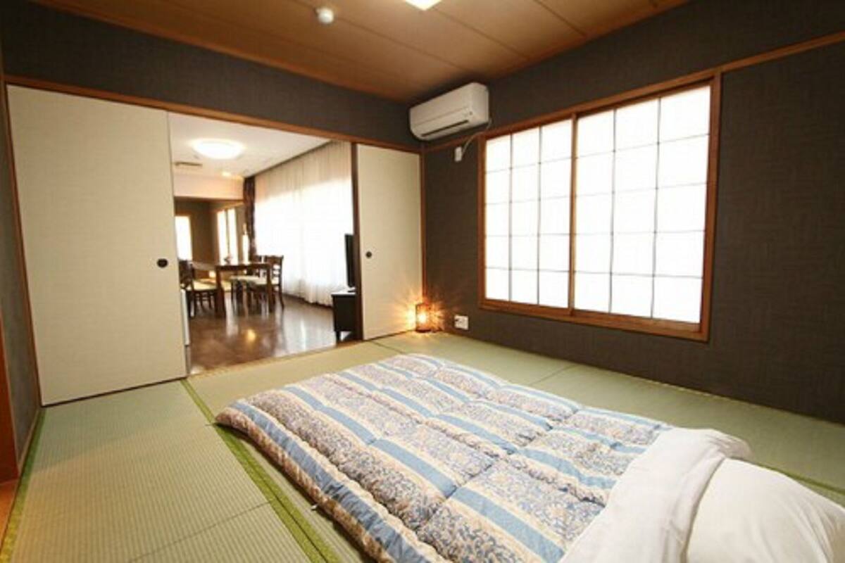 A traditional Japanese room with tatami flooring, a futon bed, sliding doors, and a large window with shoji screens.