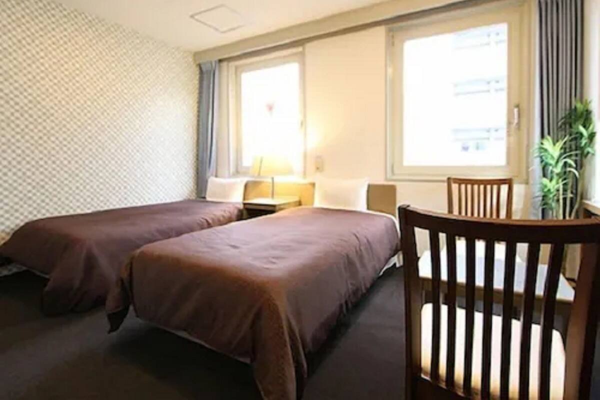 A hotel room with two beds, a desk, a chair, a window with curtains, and a plant.