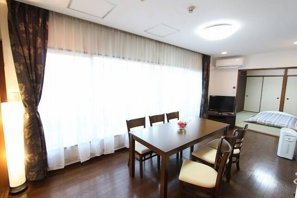 A hotel room with a wooden dining table, four chairs, a TV, a bed with a striped blanket, and a window with sheer curtains.