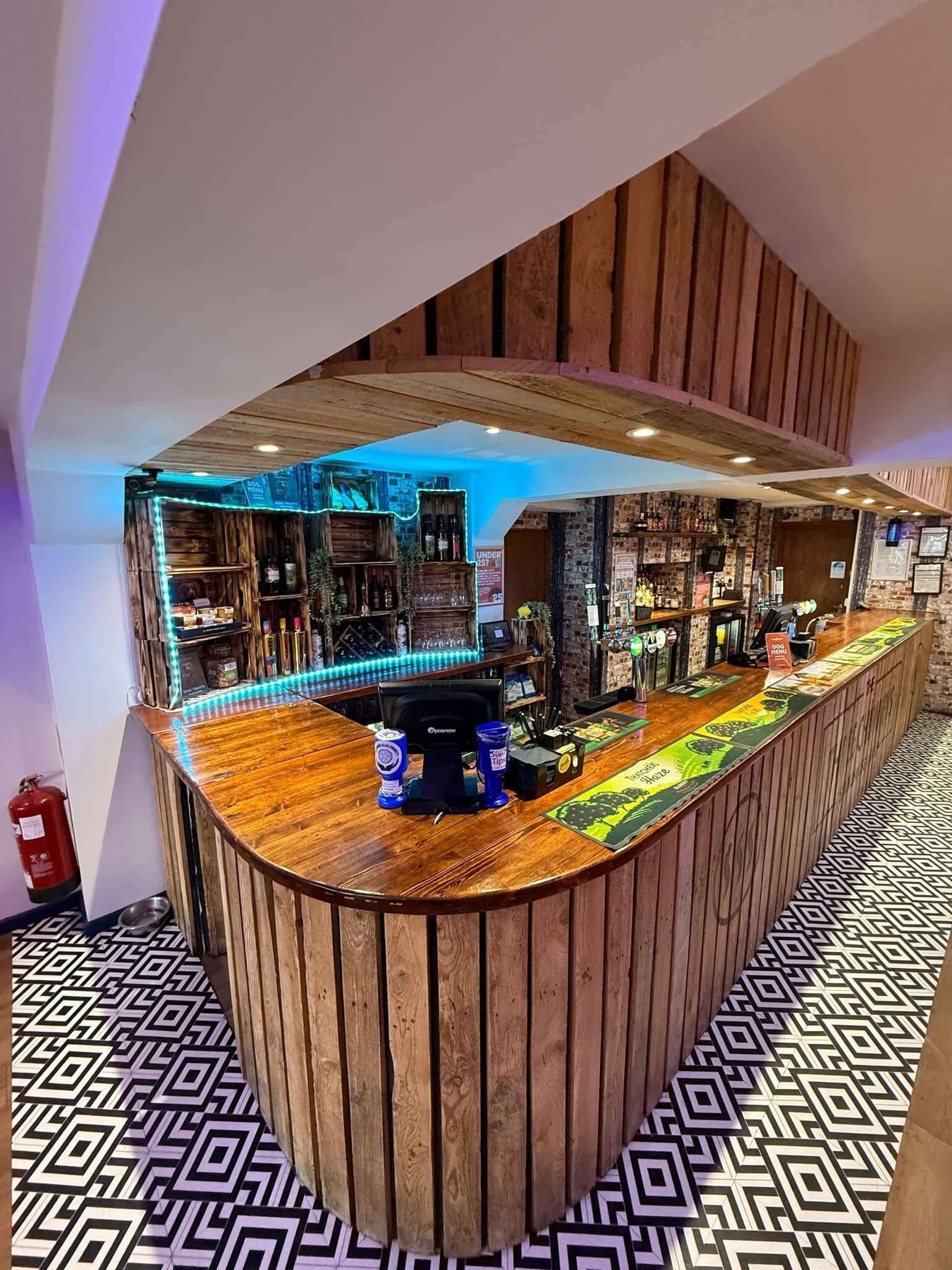 Zona de bar con mostrador de madera curvo, estanterías con botellas y suelo estampado.