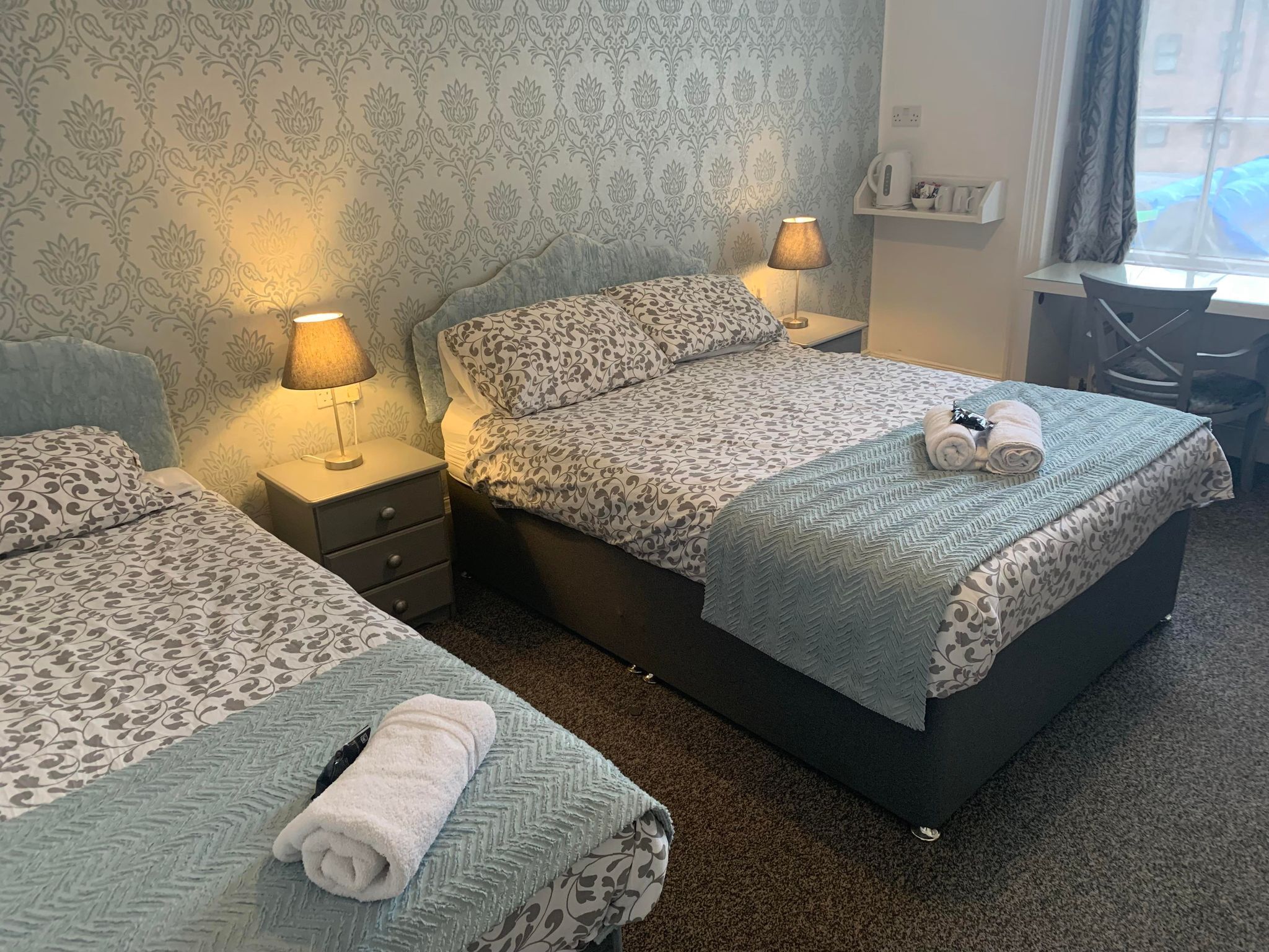 A hotel room with two beds, each with patterned bedding and a nightstand with a lamp. There is a desk with a chair and a window with curtains.
