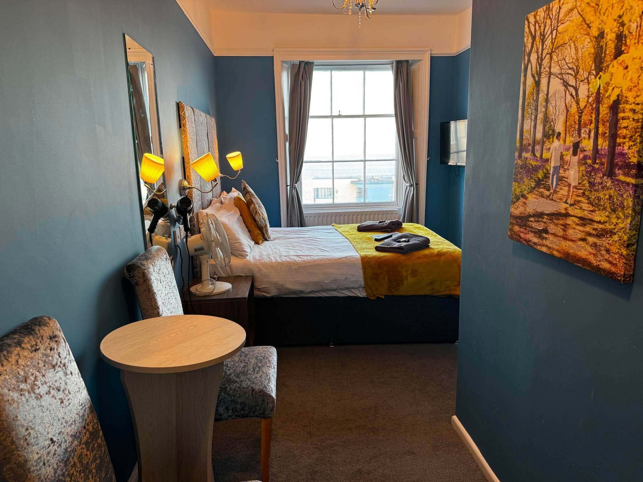 A hotel room with a bed, a round table, a chair, a large window, and a painting on the wall.